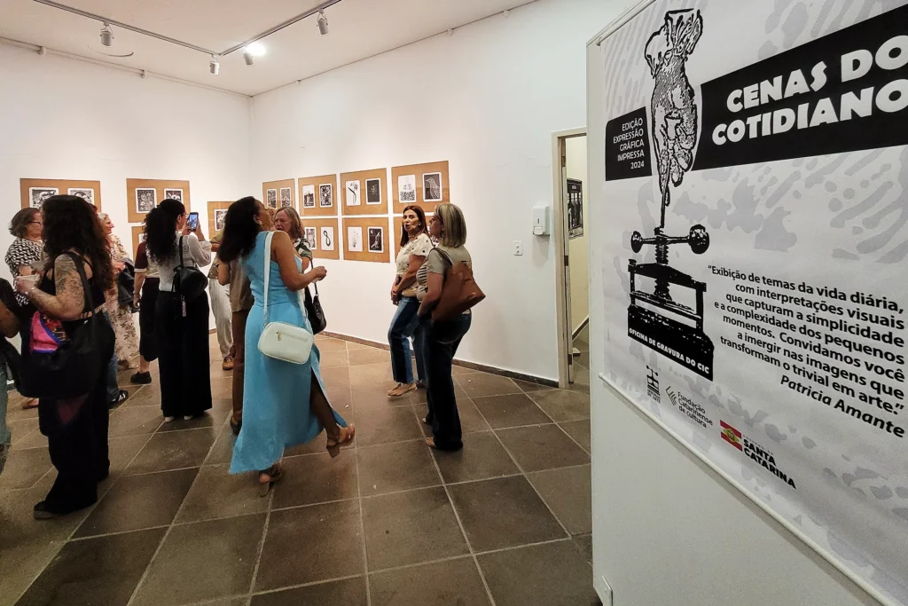 Sala da exposição com banner em primeiro plano e visitantes e obras ao fundo