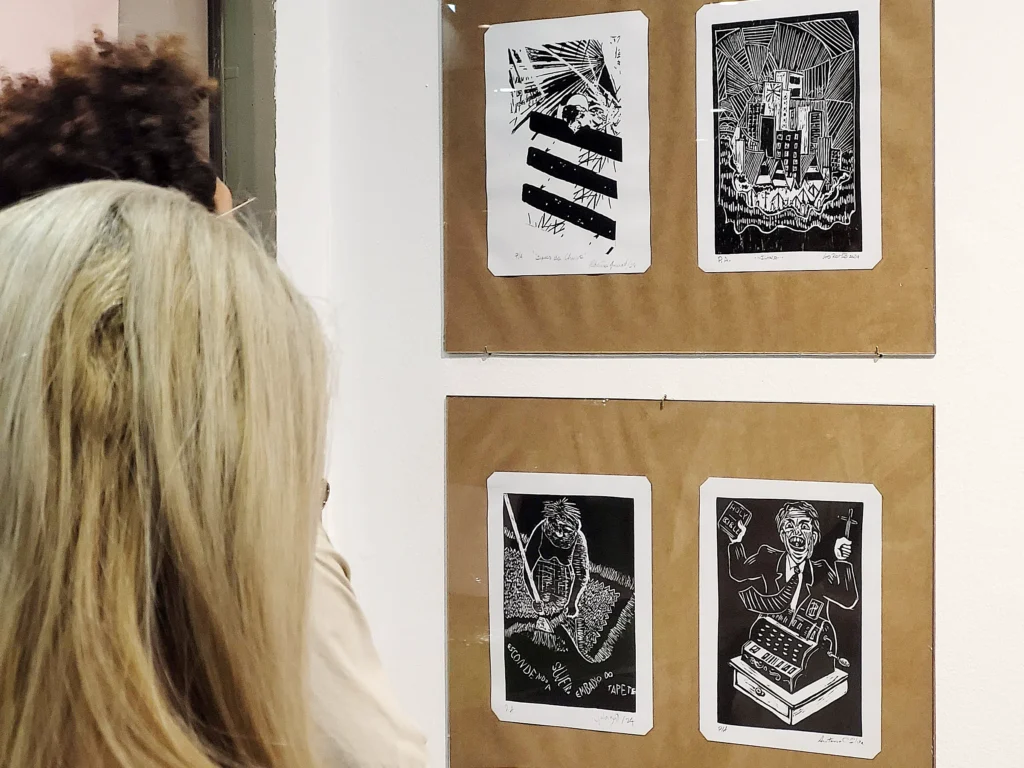 Foto com a cabeça de duas pessoas, de costas, só se veem os cabelos, observando quatro obras da exposição. As duas de baixo são de Julia Iguti e Antônio C. Silva. A de Julia mostra uma mulher varrendo sujeira para debaixo do tapete com uma vassoura